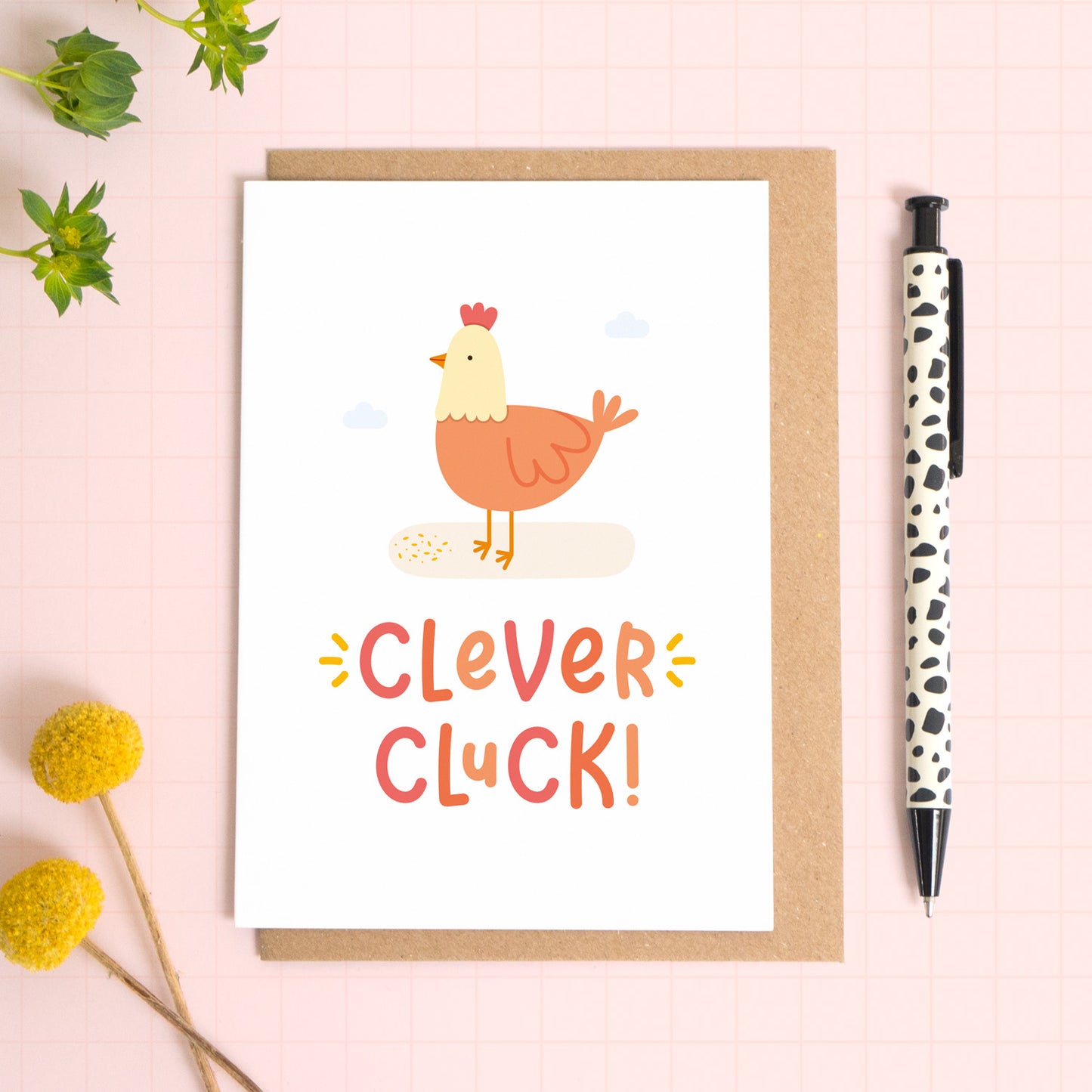 A clever cluck greeting card featuring an illustration of a chicken. This card has been photographed on top of the Kraft brown envelope that it comes with and on top of a pink background. There is foliage to the left and a pen on the right for scale.