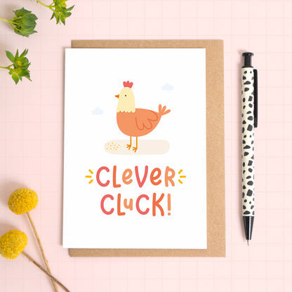 A clever cluck greeting card featuring an illustration of a chicken. This card has been photographed on top of the Kraft brown envelope that it comes with and on top of a pink background. There is foliage to the left and a pen on the right for scale.