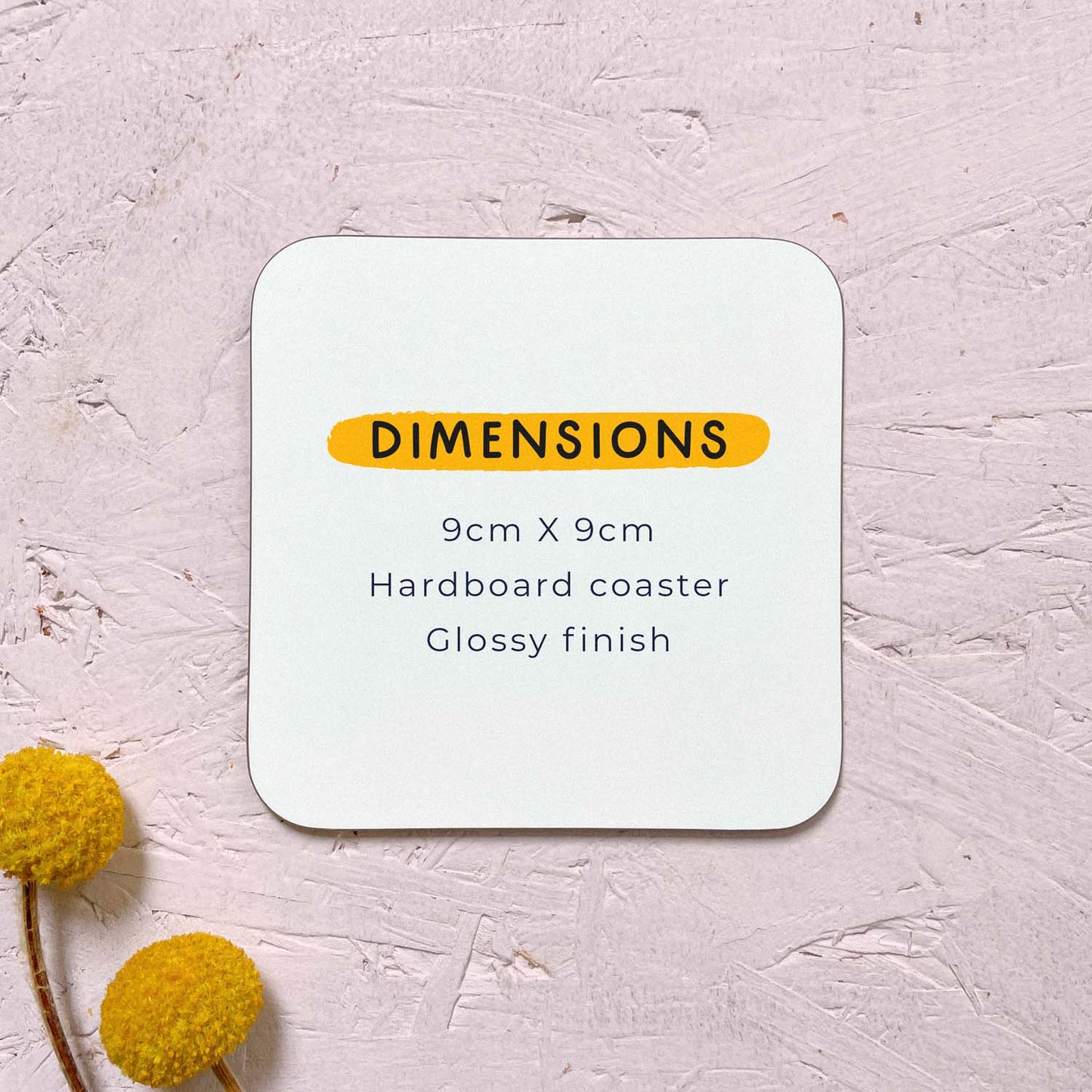 A plain coaster showing the dimensions and finish of the coaster. Each coaster is 9cm x 9cm, printed on a hardboard surface and is glossy in finish. This coaster has been photographed on a pink surface with two yellow flowers in the bottom left corner.