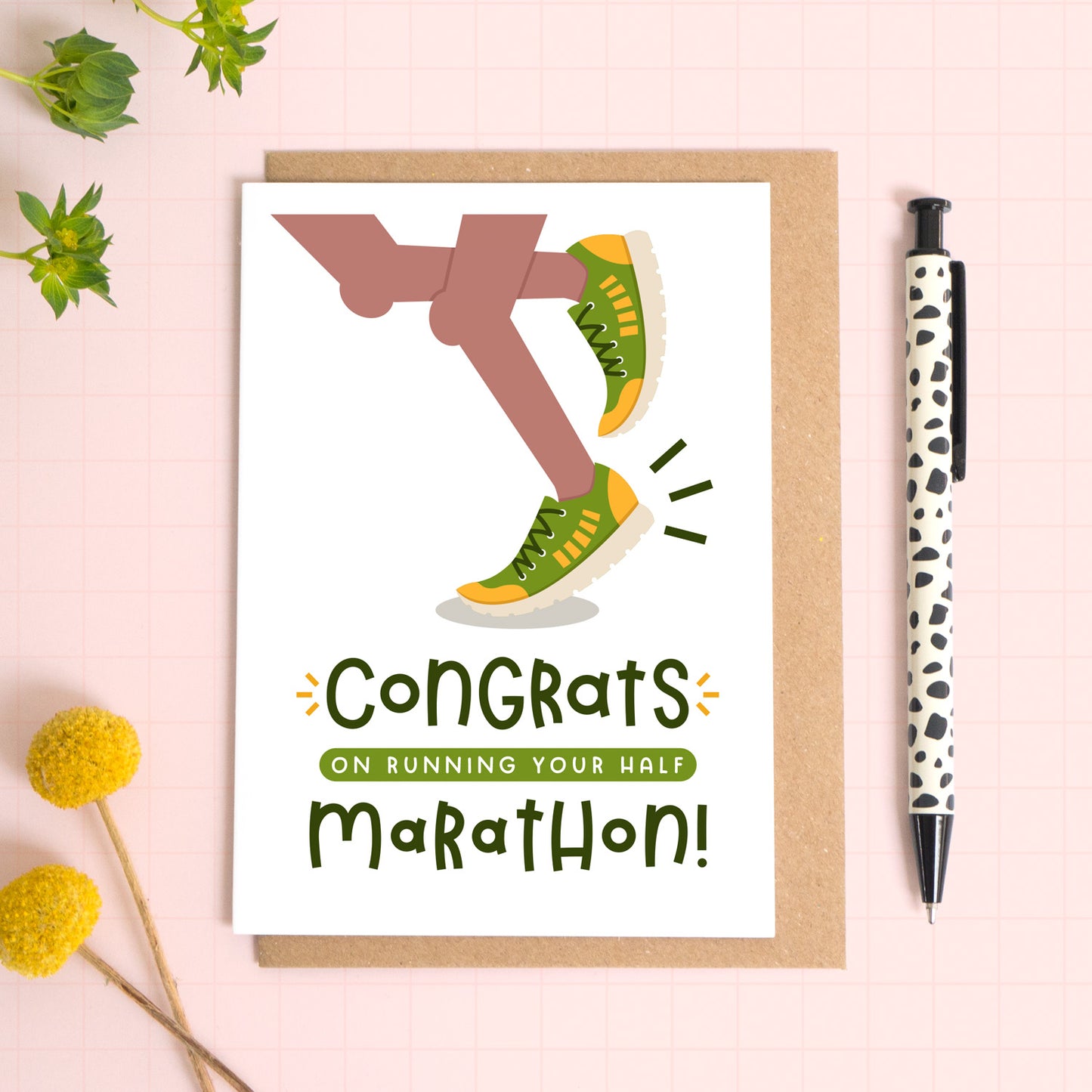 A half marathon congratulations card photographed on top of a kraft brown envelope set on a pink background surrounded by foliage and a pen for scale. The card reads 'congrats on running your half marathon' and features a pair of brown legs.