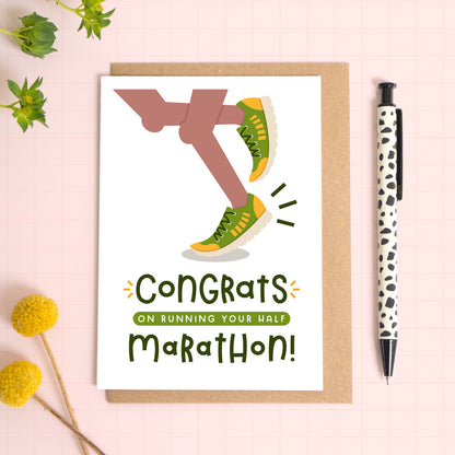 A half marathon congratulations card photographed on top of a kraft brown envelope set on a pink background surrounded by foliage and a pen for scale. The card reads 'congrats on running your half marathon' and features a pair of brown legs.