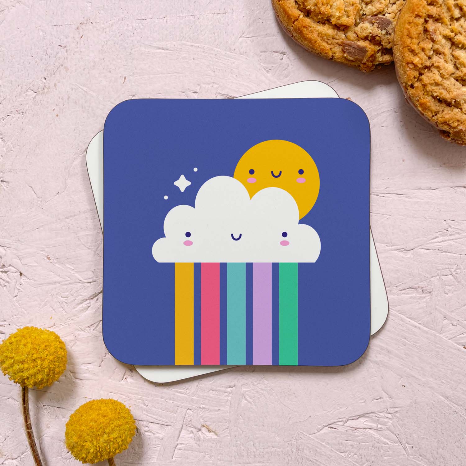 A cloud and rainbow themed coaster that has been photographed stacked on top of a blank coaster on a pink background. To the top right are two cookies and in the bottom left are two flowers. The kawaii weather themed design features a happy sun, smiling cloud and rainbow.