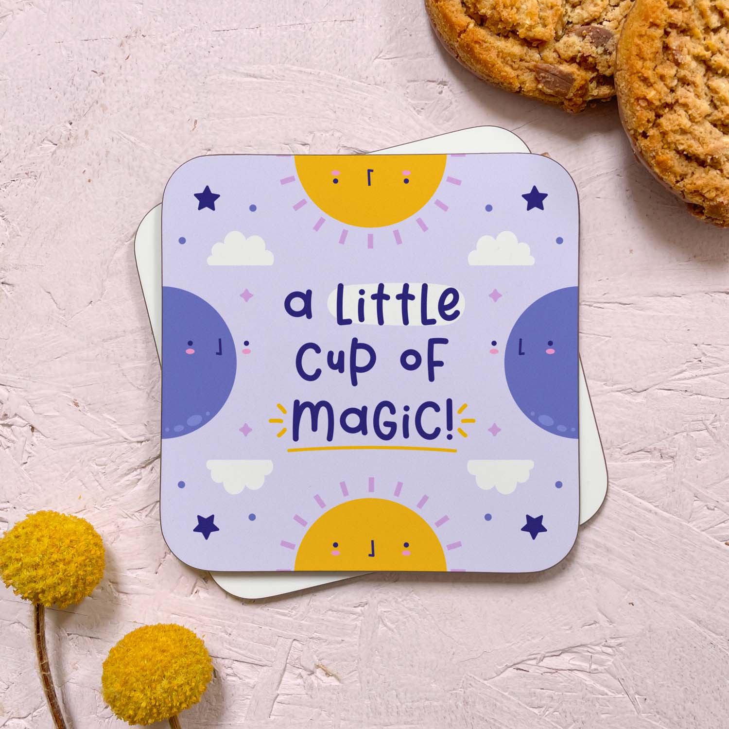 A celestial themed coaster that has been photographed stacked on top of a blank coaster on a pink background. To the top right are two cookies and in the bottom left are two flowers. The celestial design features two suns, two moons, stars and clouds with the text 'a little cup of magic!'