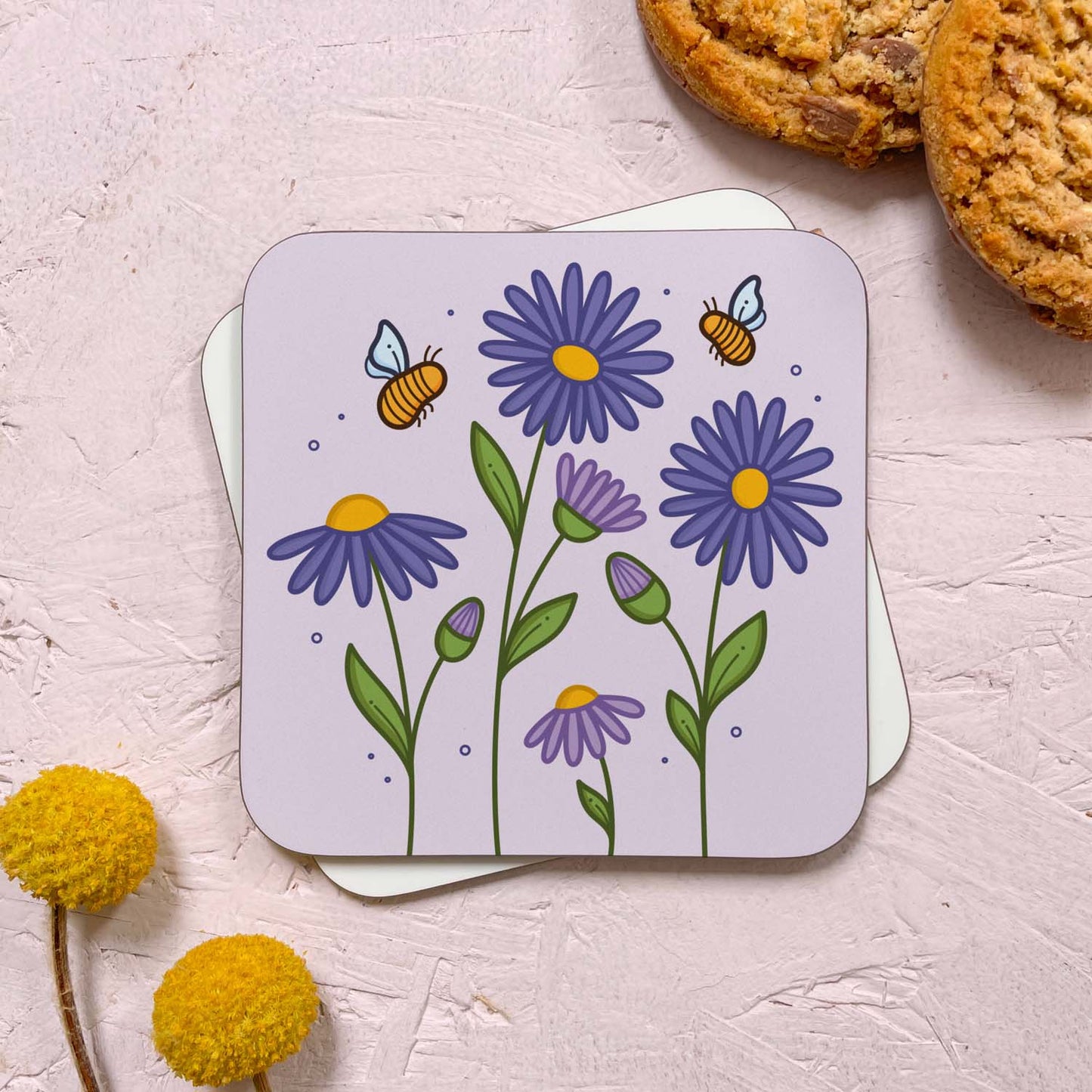 A floral themed coaster that has been photographed stacked on top of a blank coaster on a pink background. To the top right are two cookies and in the bottom left are two flowers. The floral design features purple aster flowers and bumble bees on a light lilac grey background.