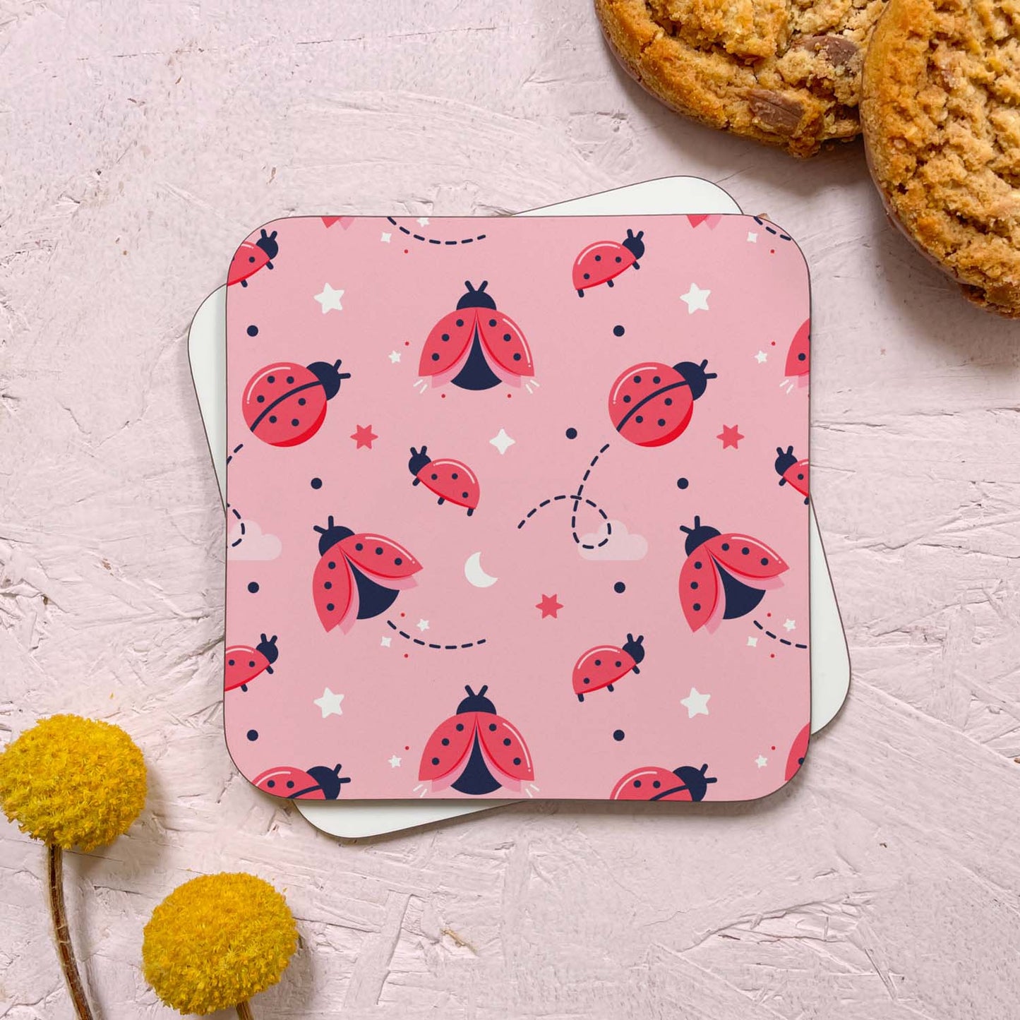 A ladybug themed coaster that has been photographed stacked on top of a blank coaster on a pink background. To the top right are two cookies and in the bottom left are two flowers. The ladybird themed design features a variety of ladybugs, stars and clouds.