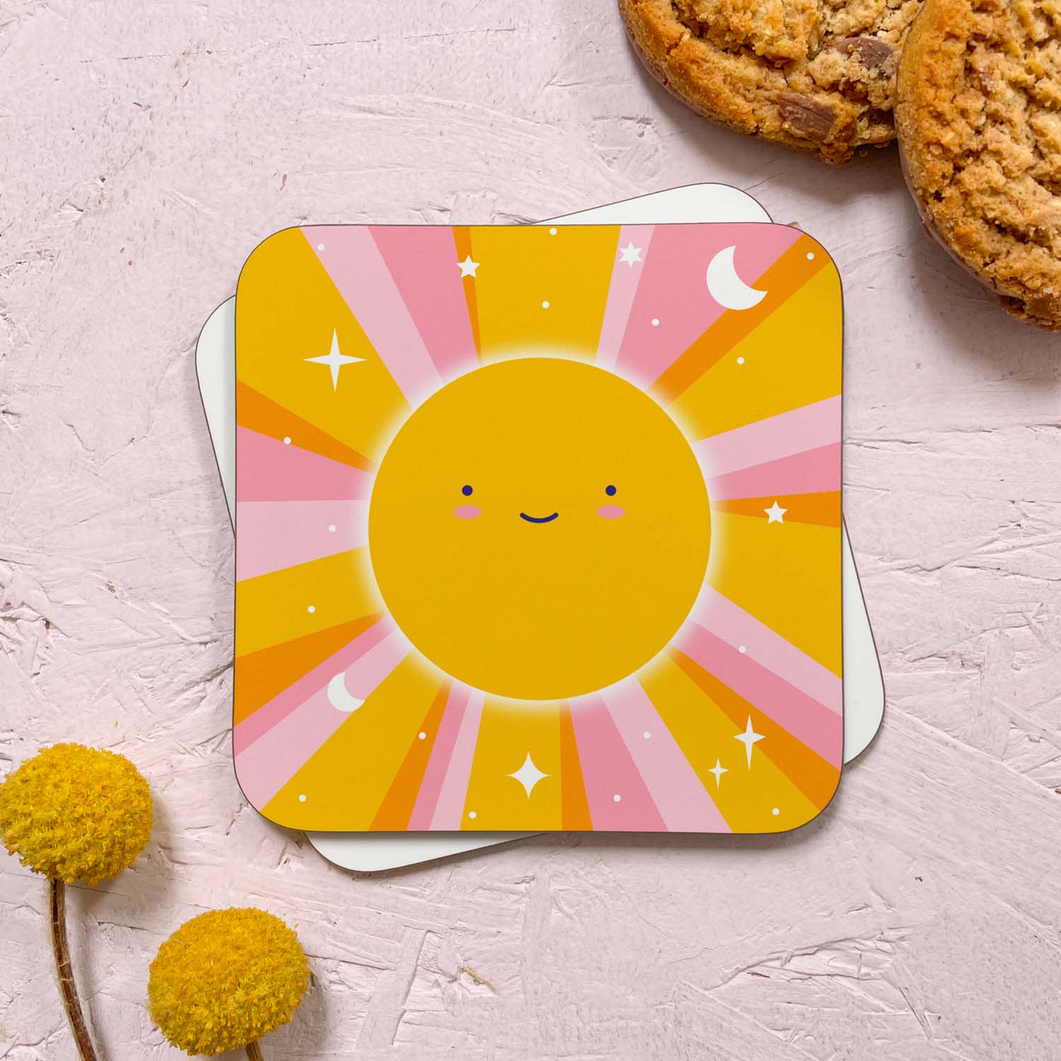 A celestial sun themed coaster that has been photographed stacked on top of a blank coaster on a pink background. To the top right are two cookies and in the bottom left are two flowers. The celestial design features a smiling sun with pink and yellow beams littered with stars, moons and dots.