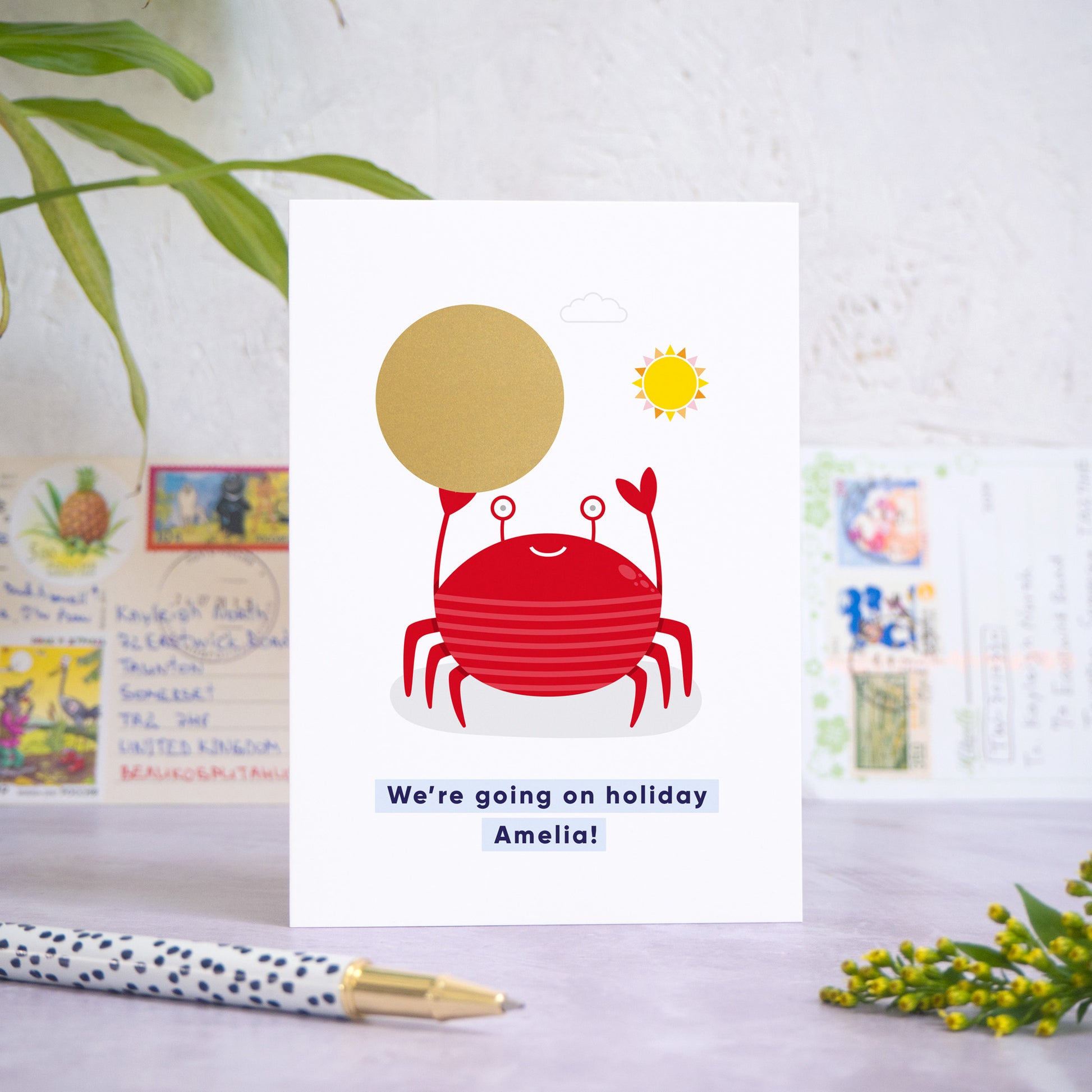 A seaside holiday reveal scratch card featuring a friendly red crab holding the gold scratch panel. The gold panel has not yet been scratched off to reveal the holiday destination. Beneath the crab illustration the text reads: “we’re going on holiday Amelia”. The card is standing on a grey surface with postcards in the background and a pen and foliage in the foreground.