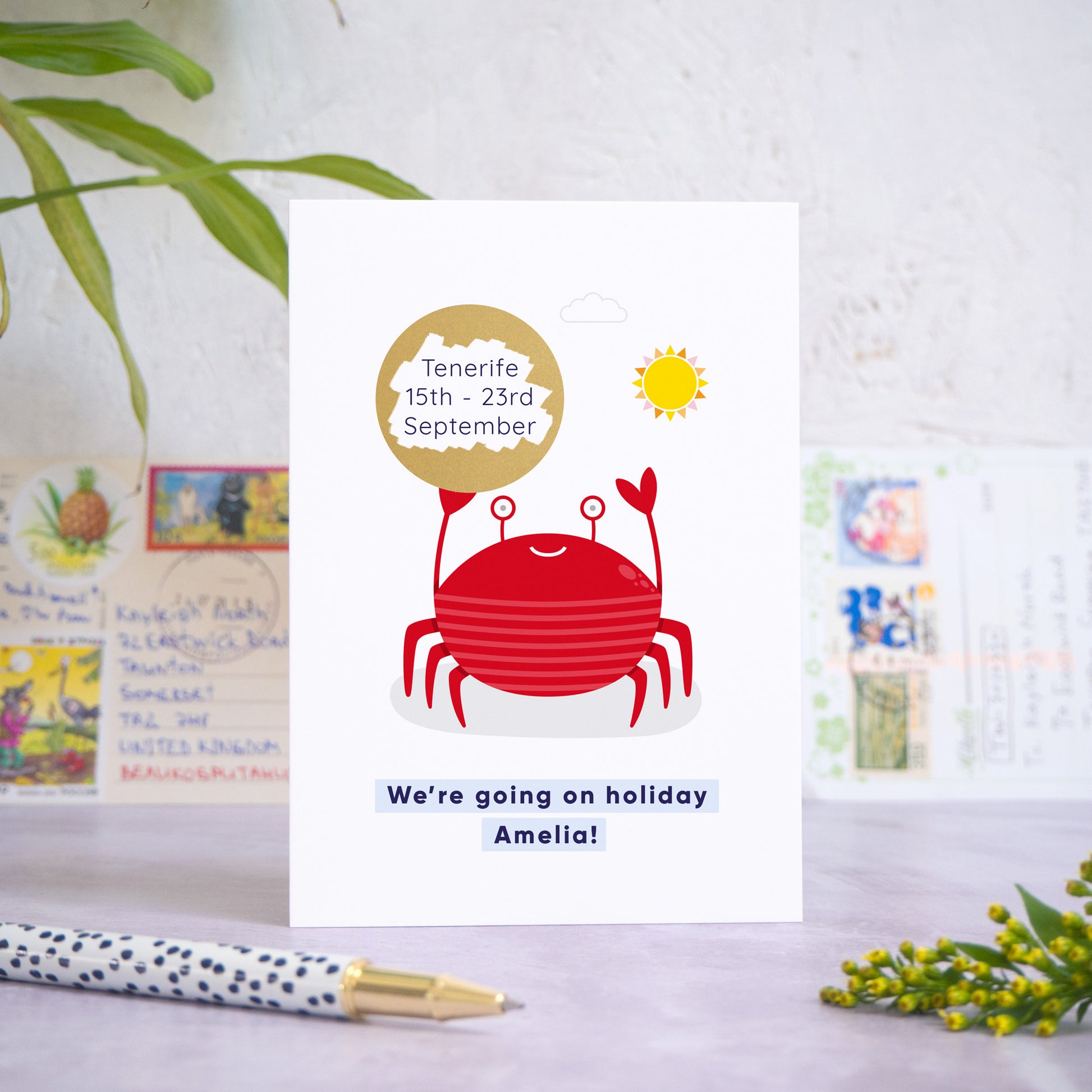 A seaside holiday reveal scratch card featuring a friendly red crab holding the gold scratch panel. The gold panel has been scratched off to reveal the holiday destination. Beneath the crab illustration the text reads: “we’re going on holiday Amelia”. The card is standing on a grey surface with postcards in the background and a pen and foliage in the foreground.