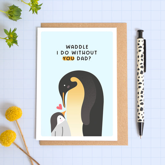 A blue card with a white border featuring two penguins and the phrase 'Waddle I do without you Dad!’. The card is laid on a kraft brown envelope on top of a blue background next to a pen and flowers.