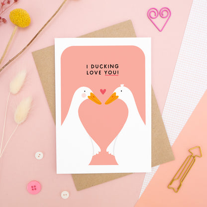 A Valentine’s pun card featuring two cute white ducks holding wings with the text ‘I Ducking Love You’, photographed on a pink background with floral props, paper clips, and buttons. 