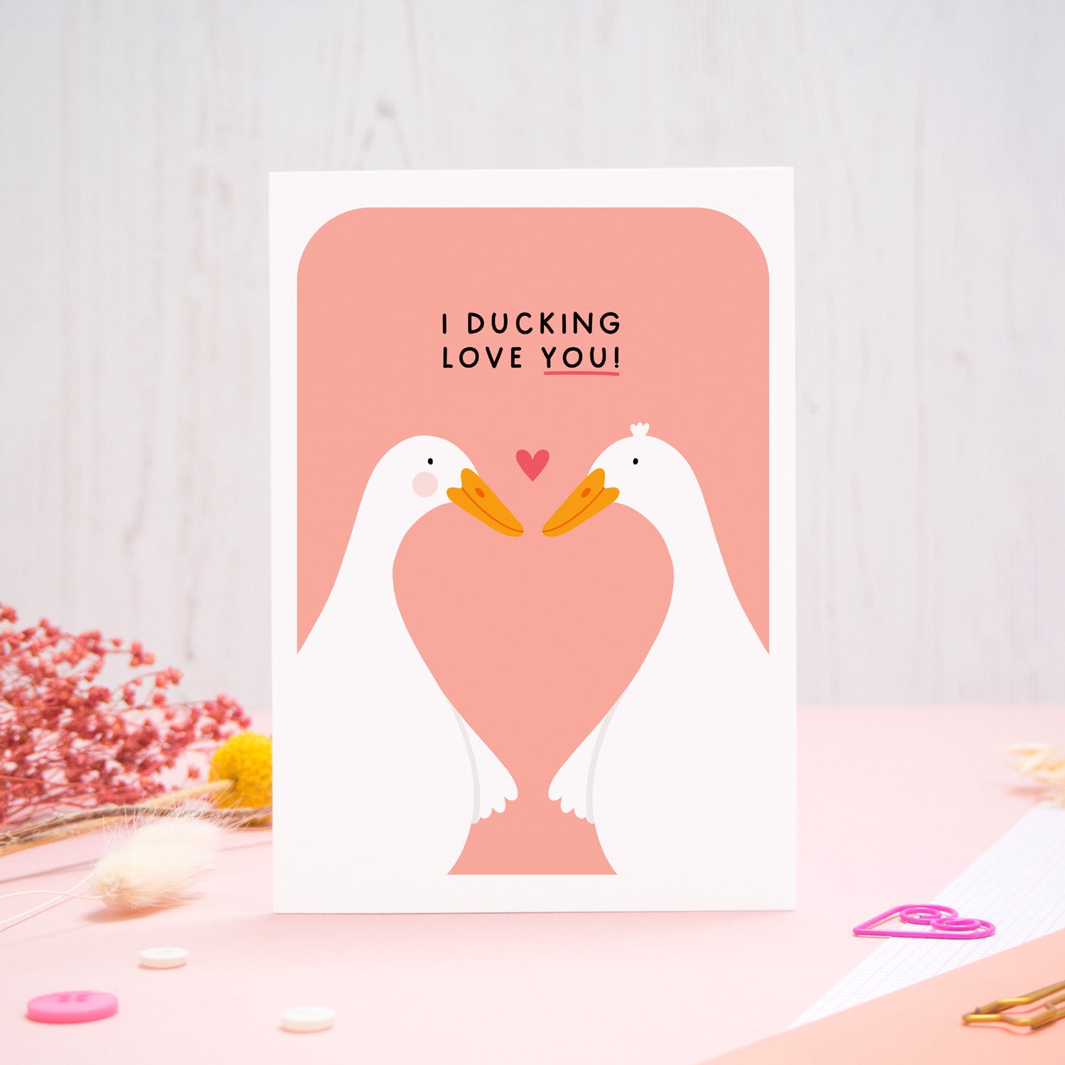 A Valentine’s pun card featuring two cute white ducks holding wings with the text ‘I Ducking Love You’, photographed stood on a pink and white background with floral props, paper clips, and buttons. 