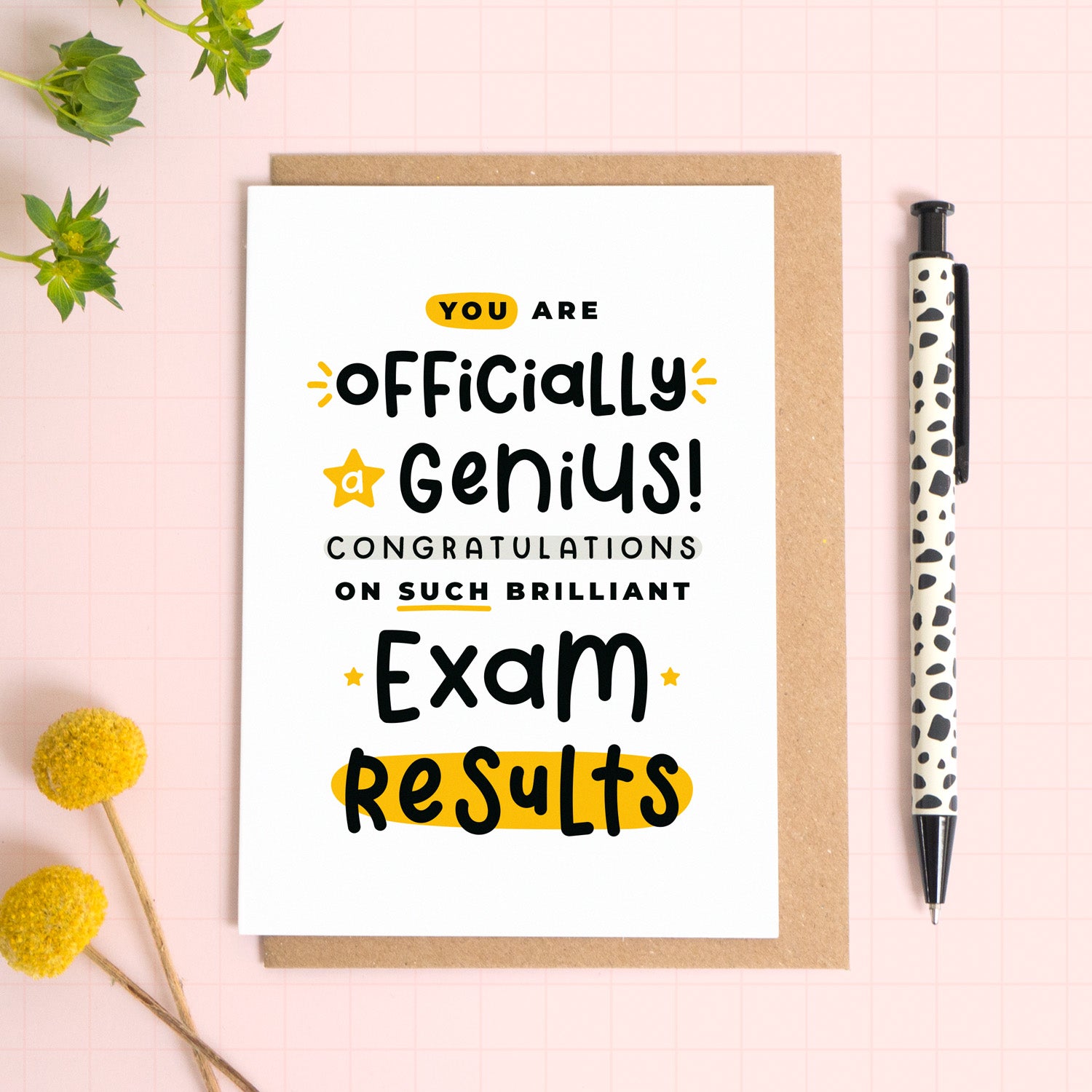 An exam results congratulations card for someone who has just received brilliant exam results. This card has been photographed on top of the Kraft brown envelope that it comes with and on top of a pink background. There is foliage to the left and a pen on the right for scale. This is the black and yellow version.