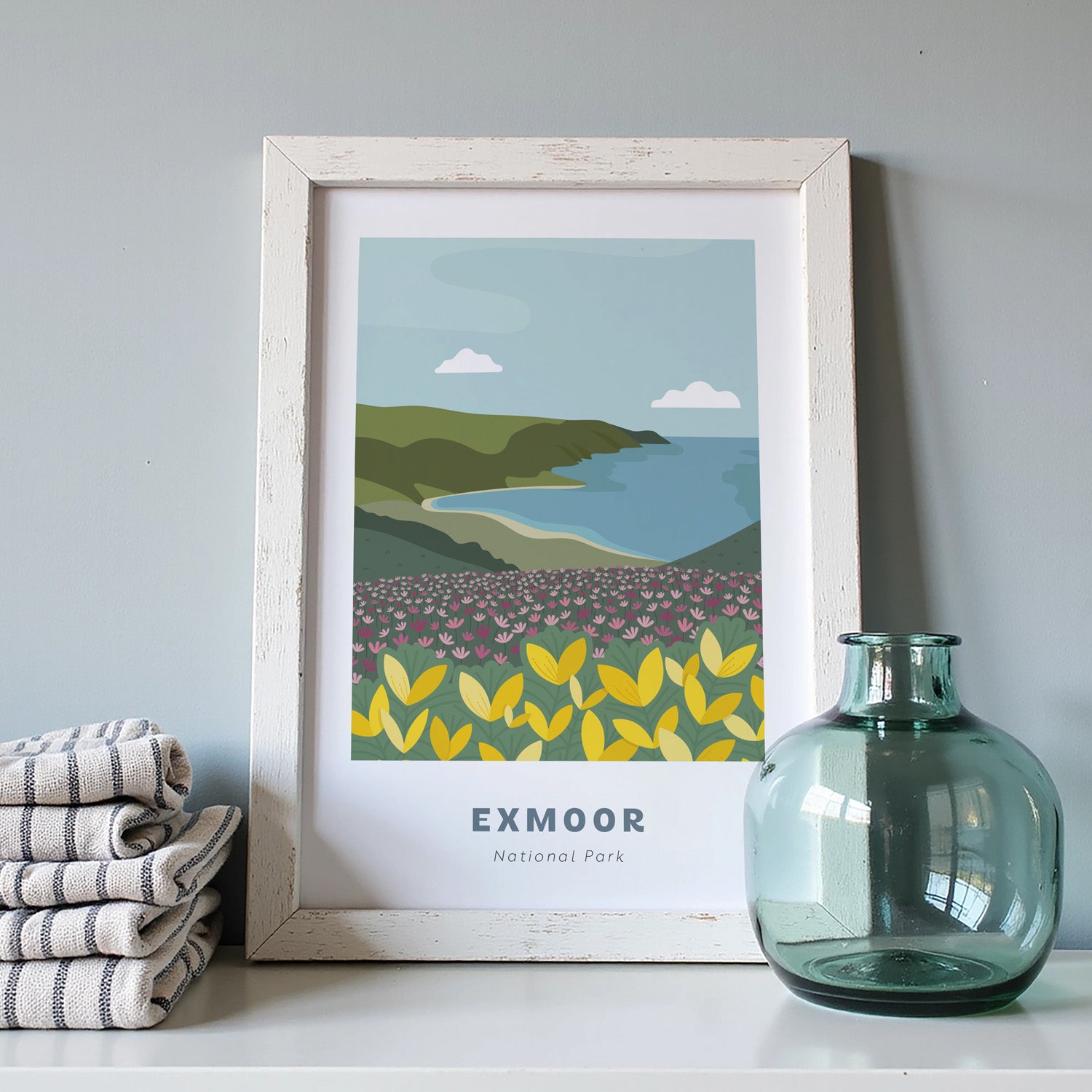 Exmoor National Park art print in a white frame on a light grey background with a pile of cloths and blue vase.