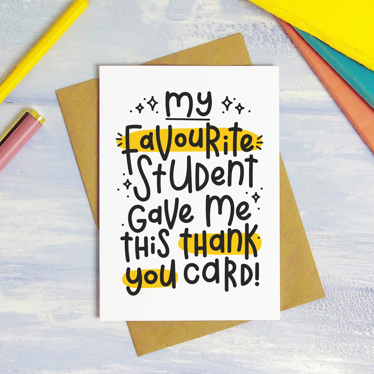 A favourite student thank you teacher card photographed on a purple surface with colourful text books in the background. The card reads "my favourite student gave me this thank you card!"