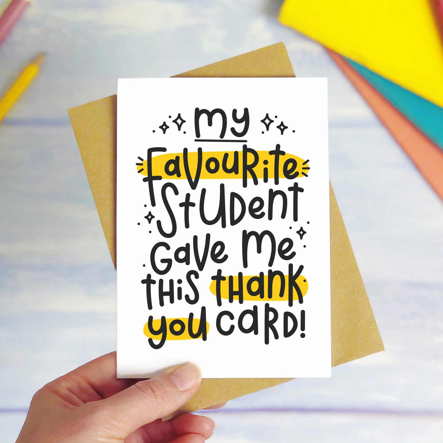 A favourite student thank you teacher card held over a purple surface with colourful text books in the background. The card reads "my favourite student gave me this thank you card!"