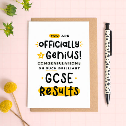 An exam results congratulations card for someone who has just received brilliant GCSE results. This card has been photographed on top of the Kraft brown envelope that it comes with and on top of a pink background. There is foliage to the left and a pen on the right for scale. This is the black and yellow version.