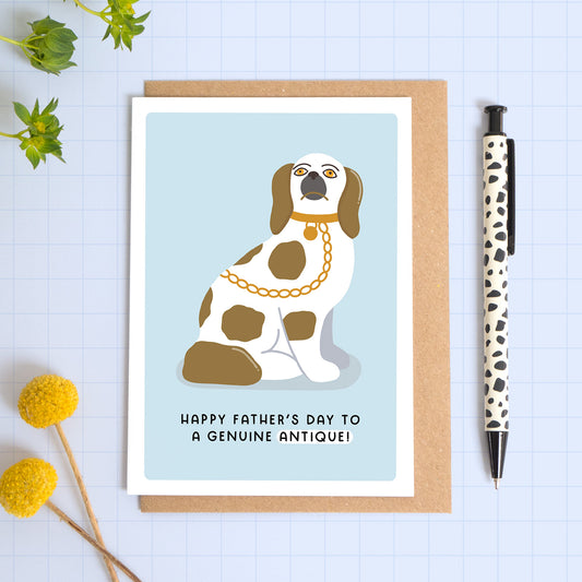 A blue card with a white border featuring a seated Staffordshire dog in the King Charles cavalier style and the phrase ‘Happy Father’s day to a genuine antique!’. The card is laid on a kraft brown envelope on top of a blue background next to a pen and flowers.