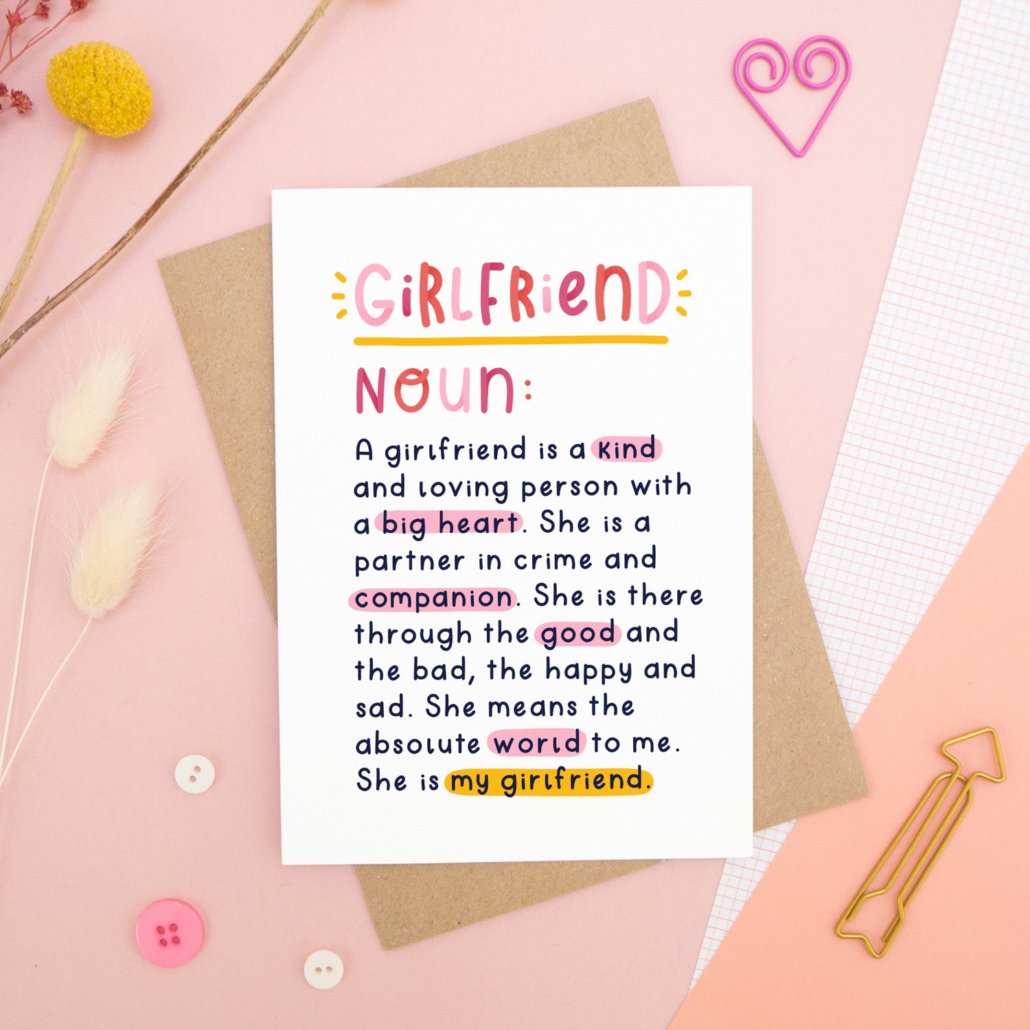 A heartfelt, colourful dictionary definition style card with the definition of a girlfriend, photographed on a pink background with floral props, paper clips, and buttons. 