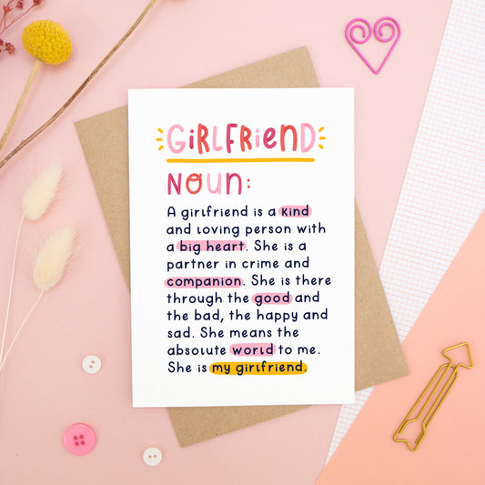 A heartfelt, colourful dictionary definition style card with the definition of a girlfriend, photographed on a pink background with floral props, paper clips, and buttons. 