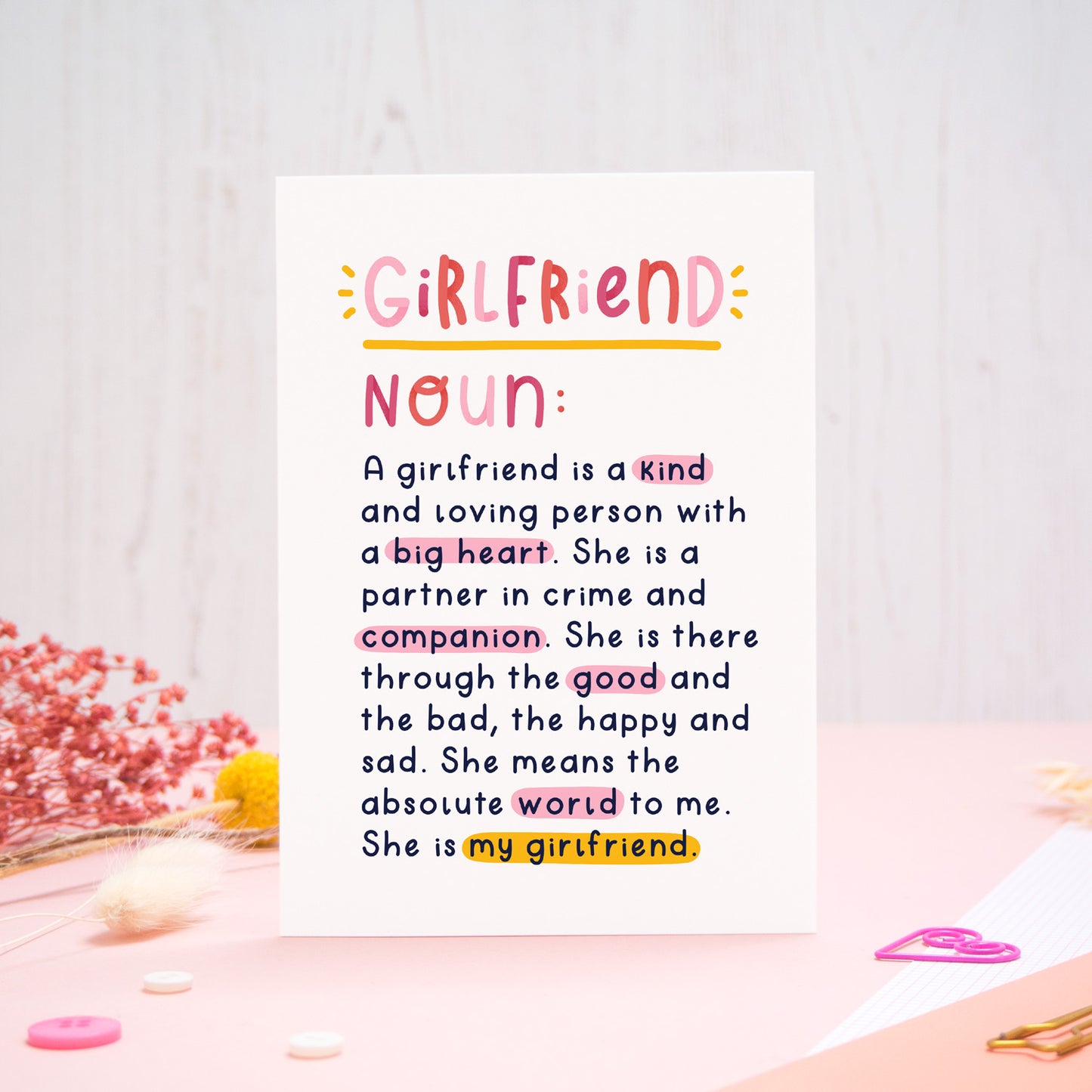 A heartfelt, colourful dictionary definition style card with the definition of a girlfriend, photographed stood on a pink and white background with floral props, paper clips, and buttons. 