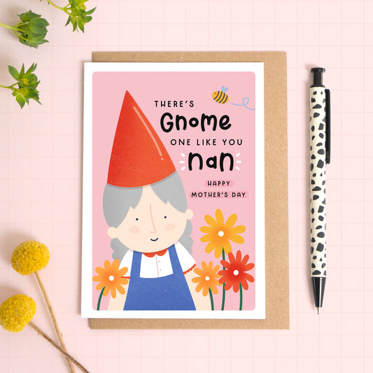 A garden gnome card with a white border featuring the phrase 'There's gnome one like you nan, happy mother's day!'. The card is laid on a kraft brown envelope on top of a pink background next to a pen and flowers.