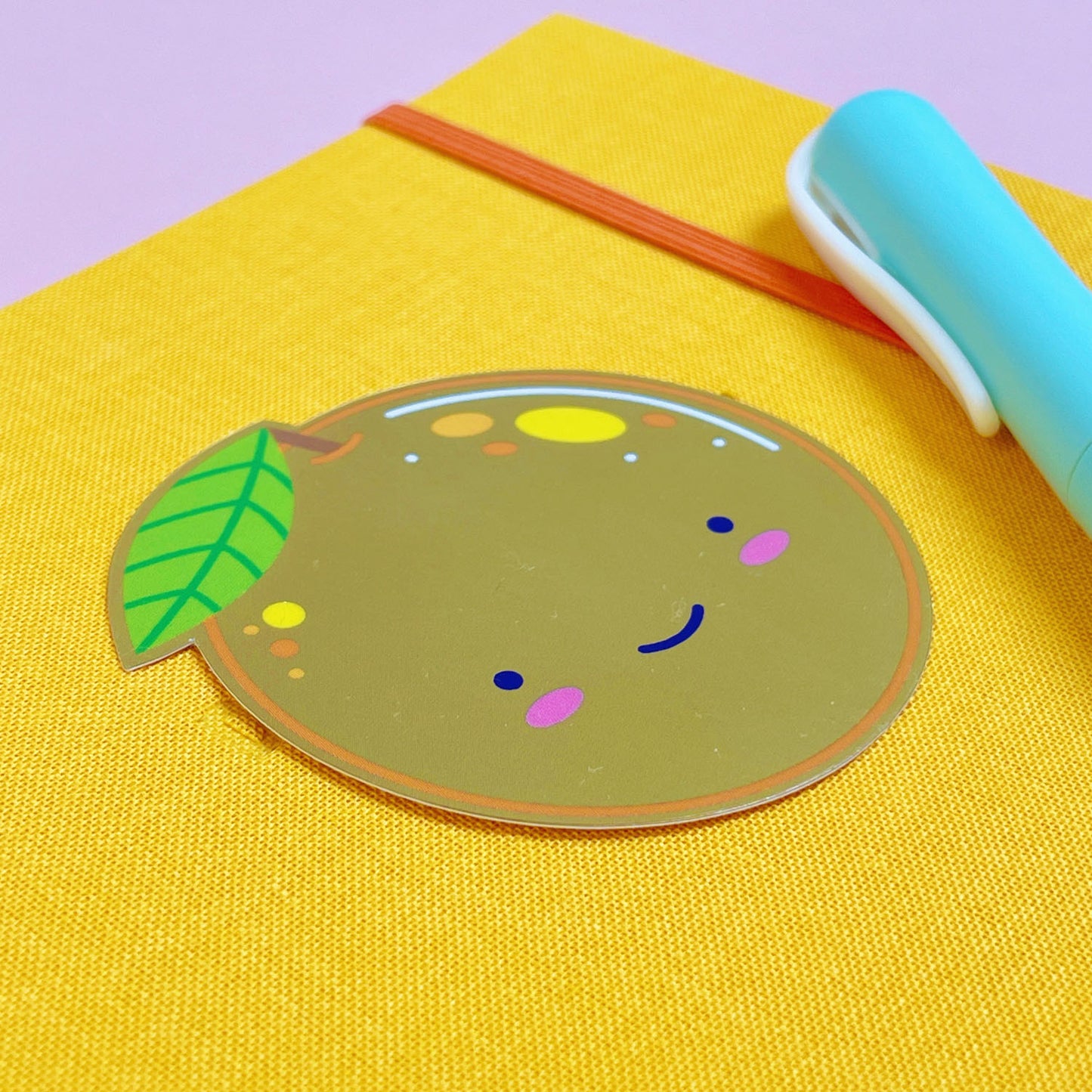 A shiny mirror effect gold sticker featuring a smiling orange lying on top of a yellow journal on a pink background