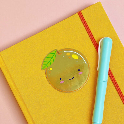A shiny mirror effect gold sticker featuring a smiling orange lying on top of a yellow journal on a pink background