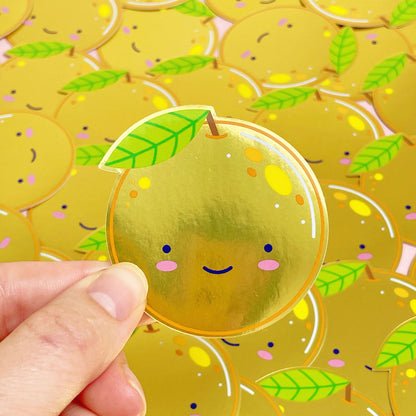 A shiny mirror effect gold sticker featuring a smiling orange being held over a pile of stickers of the same design