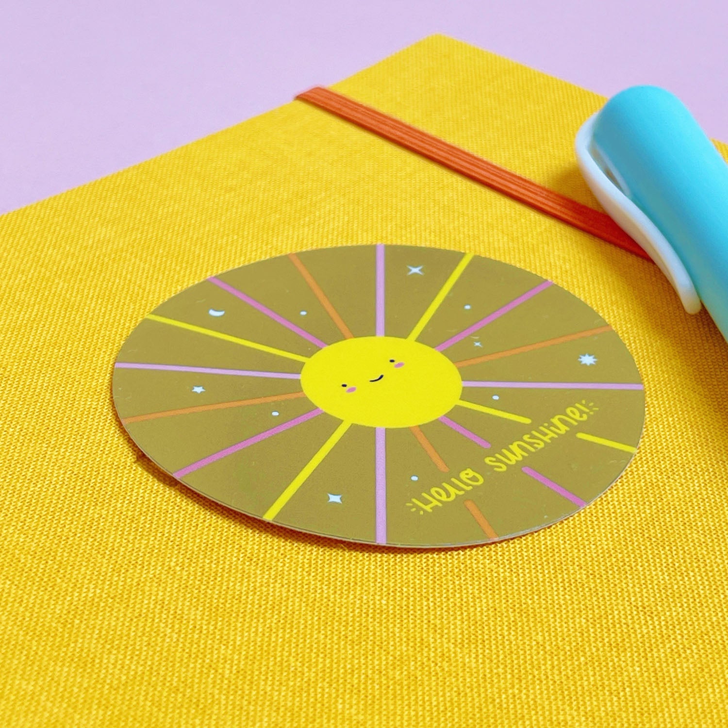 A shiny mirror effect gold sticker featuring a smiling sun lying on top of a yellow journal on a pink background