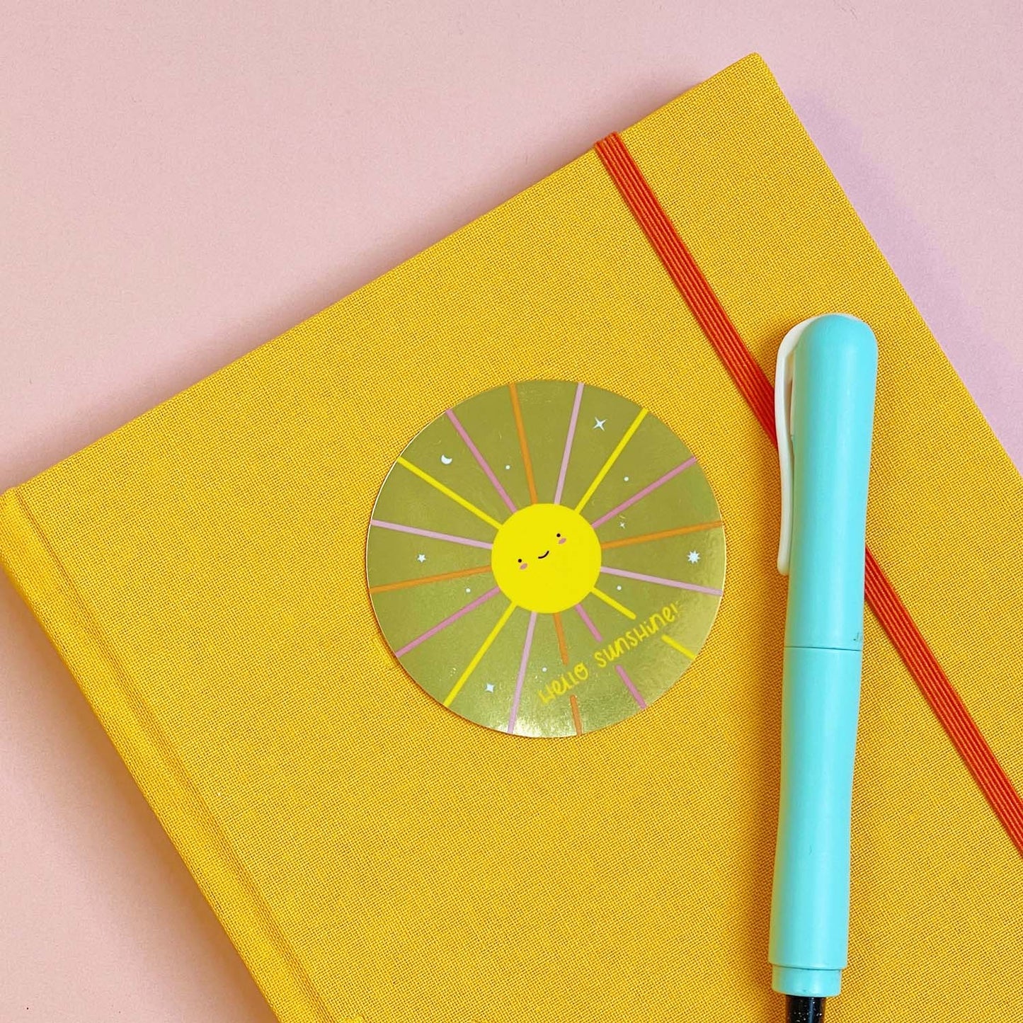 A shiny mirror effect gold sticker featuring a smiling sun lying on top of a yellow journal on a pink background