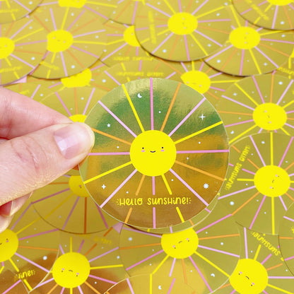 A shiny mirror effect gold sticker featuring a smiling sun being held over a pile of stickers of the same design