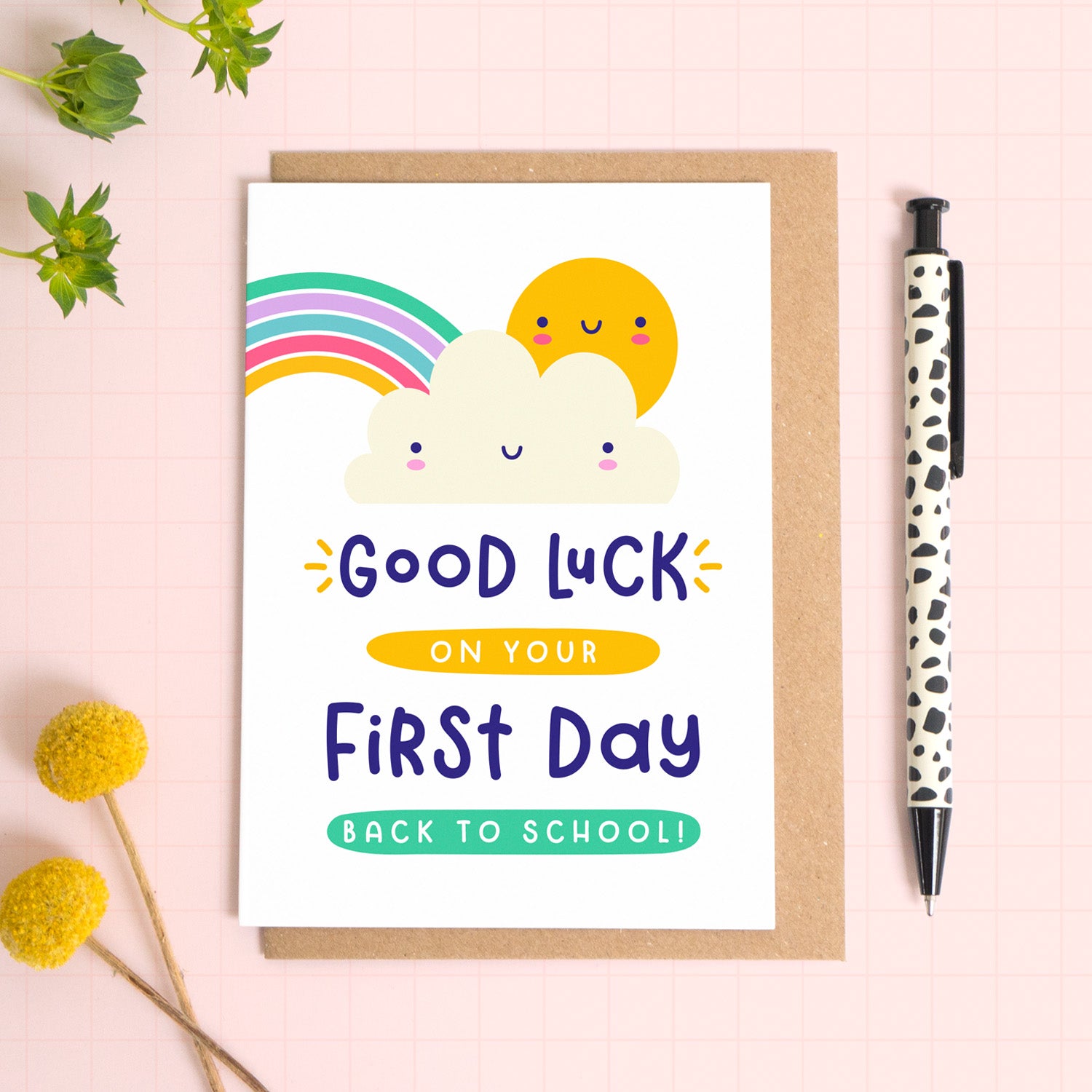 A good luck on your first day back to school card featuring a rainbow, cute cloud and happy sun. The card has been photographed on it's kraft brown envelope on a pink surface with foliage and a pen for scale. 