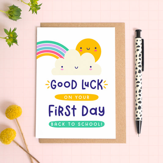 A good luck on your first day back to school card featuring a rainbow, cute cloud and happy sun. The card has been photographed on it's kraft brown envelope on a pink surface with foliage and a pen for scale. 