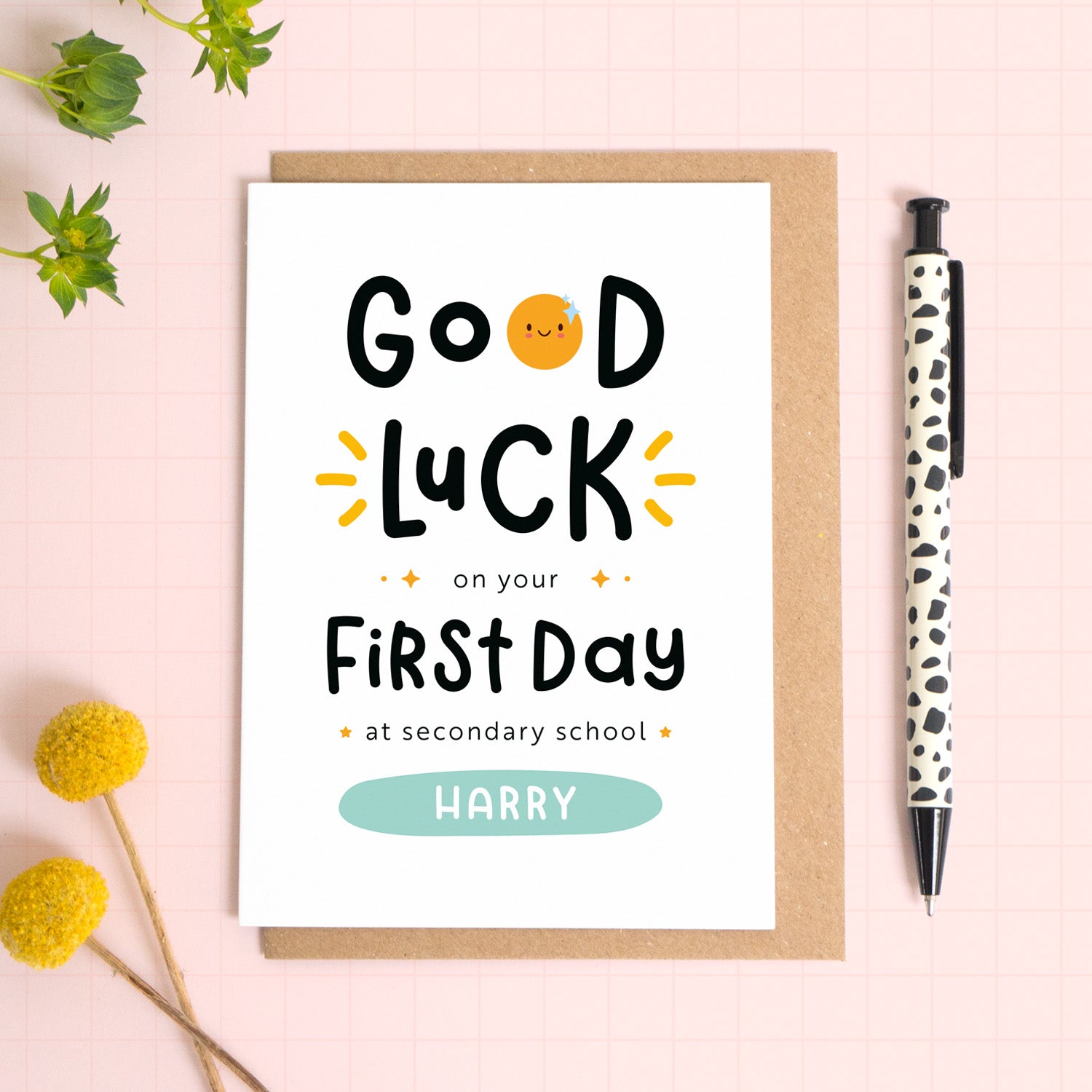 A good luck on your first day at secondary school card personalised with the recipients name. The card has been photographed on it's kraft brown envelope on a pink surface with foliage and a pen for scale. This is the blue version of the card.