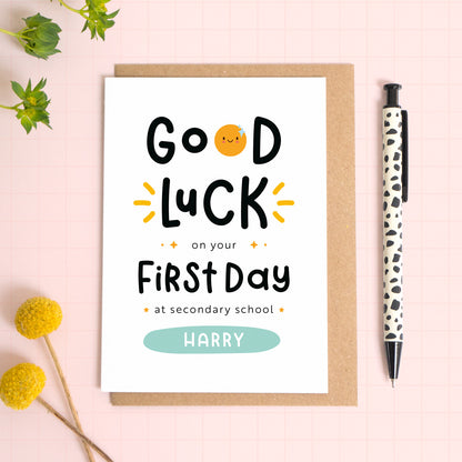 A good luck on your first day at secondary school card personalised with the recipients name. The card has been photographed on it's kraft brown envelope on a pink surface with foliage and a pen for scale. This is the blue version of the card.