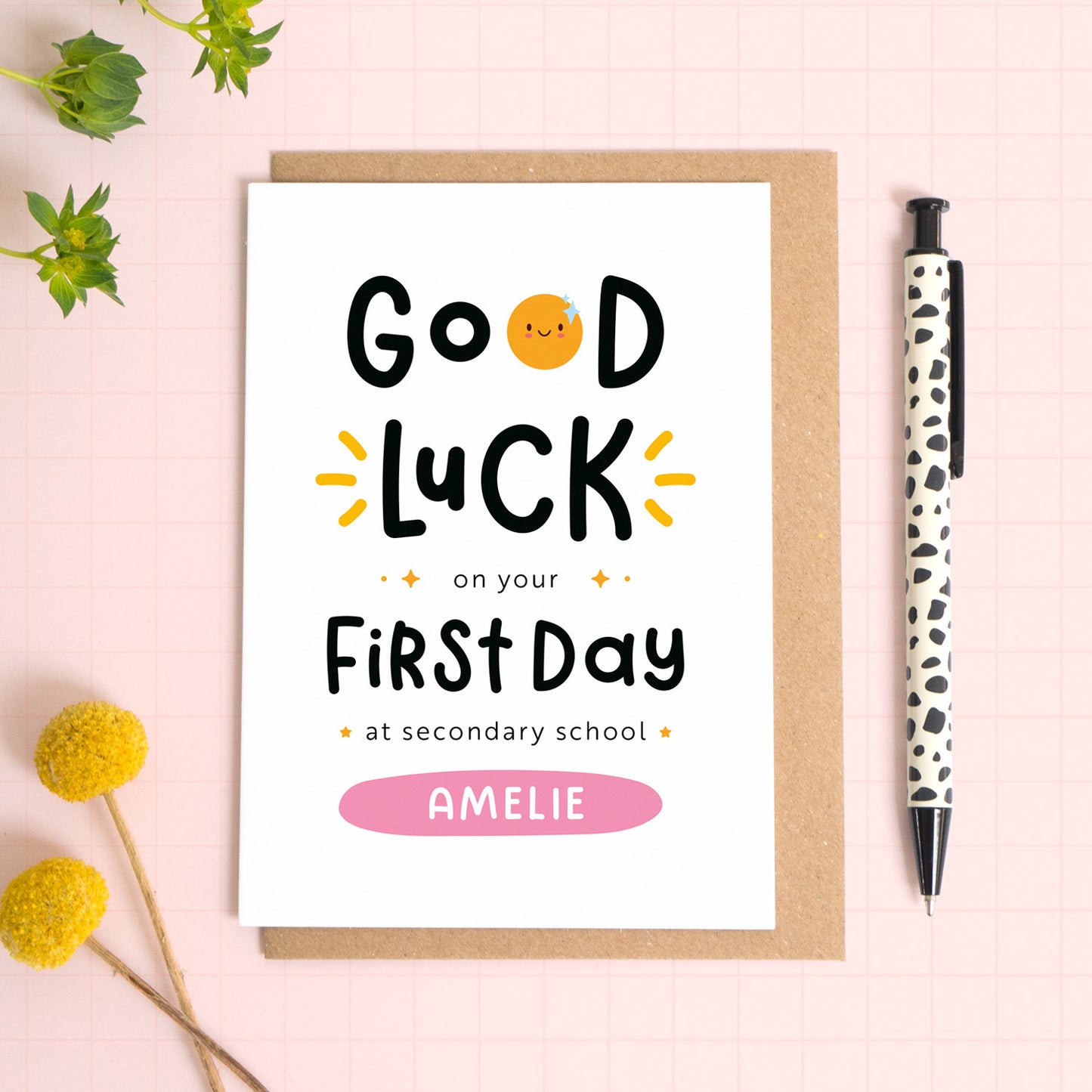 A good luck on your first day at secondary school card personalised with the recipients name. The card has been photographed on it's kraft brown envelope on a pink surface with foliage and a pen for scale. This is the pink version of the card.