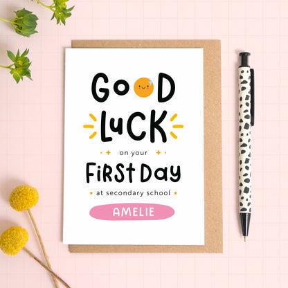 A good luck on your first day at secondary school card personalised with the recipients name. The card has been photographed on it's kraft brown envelope on a pink surface with foliage and a pen for scale. This is the pink version of the card.