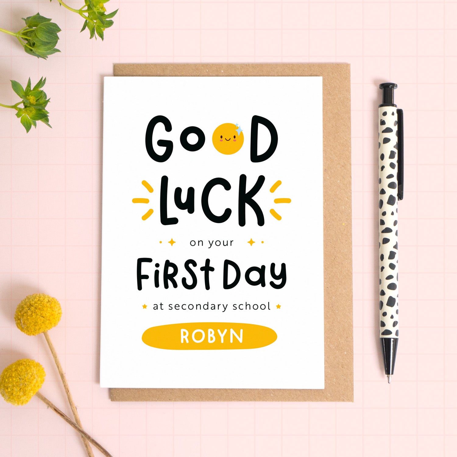 A good luck on your first day at secondary school card personalised with the recipients name. The card has been photographed on it's kraft brown envelope on a pink surface with foliage and a pen for scale. This is the yellow version of the card.