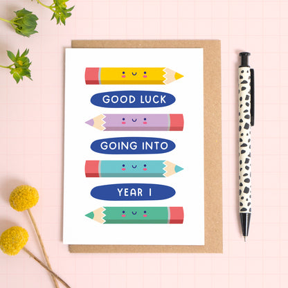 A good luck going to year 1 back to school card featuring a lots of colourful pencils. The card has been photographed on it's kraft brown envelope on a pink surface with foliage and a pen for scale. 