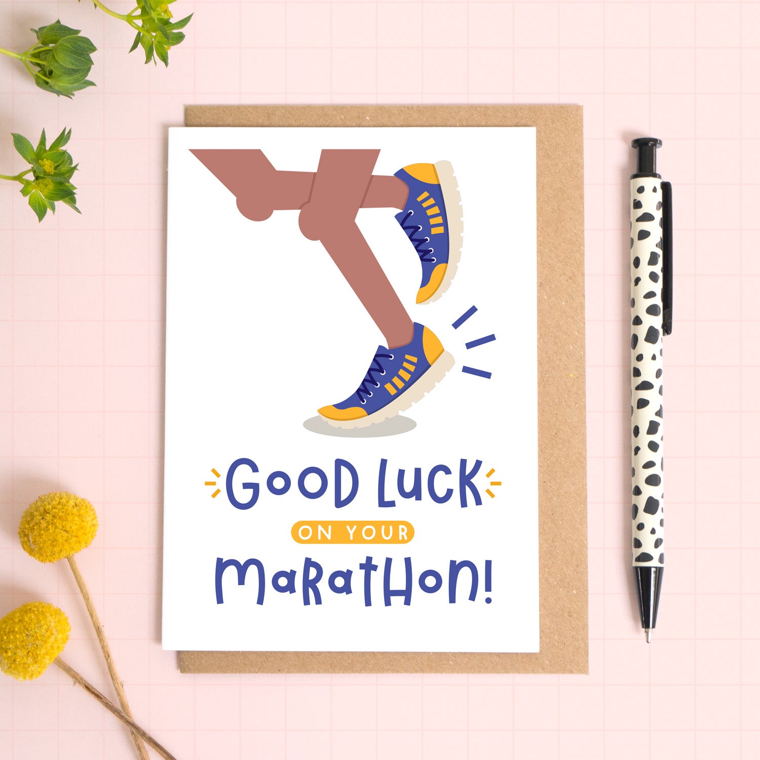 A marathon good luck card photographed on top of a kraft brown envelope set on a pink background surrounded by foliage and a pen for scale. The card reads 'good luck on your marathon' and features a pair of brown legs.