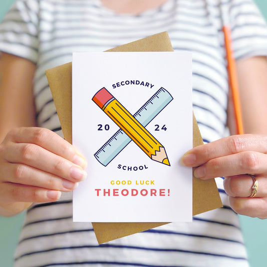Good luck for secondary personalised back to school card featuring a pencil, ruler and the young persons name. The card is being held by a person in a stripy white and blue t-shirt.