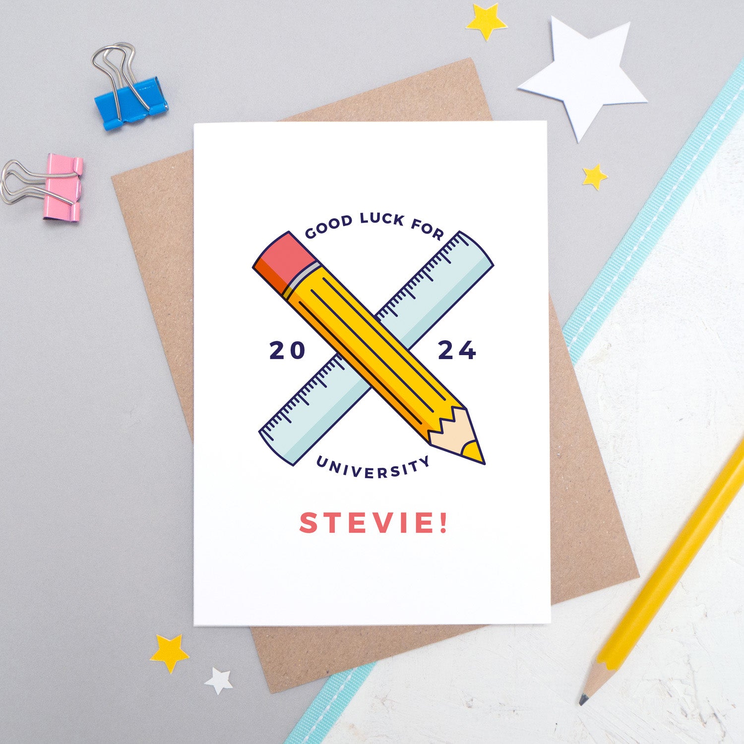 A good luck for university, personalised back to school card personalised with a young persons name, and a pencil and ruler illustration. The card is laid on it's kraft brown envelope on a grey and white background.