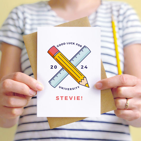 Good luck for university personalised back to school card featuring a pencil, ruler and the young persons name. The card is being held by a person in a stripy white and blue t-shirt.
