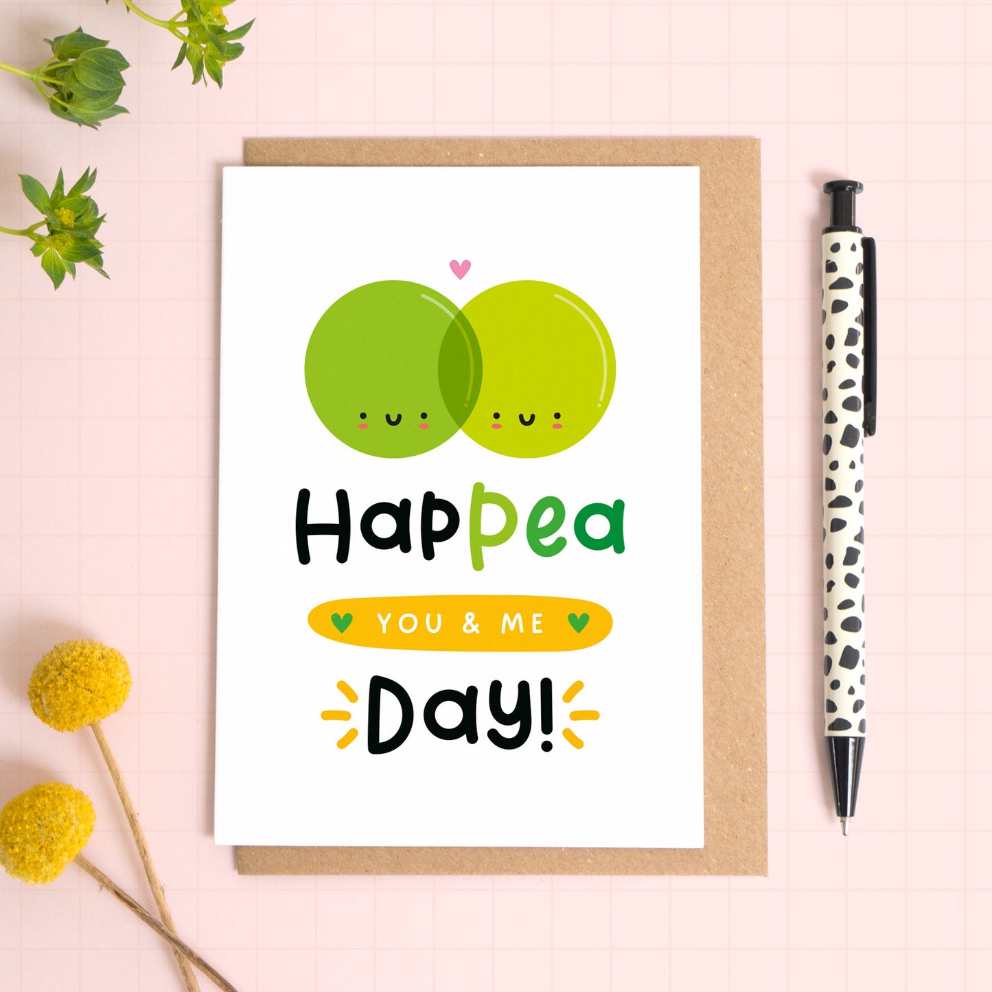 A cute anniversary card with two smiling, overlapping peas with the text ‘happea you & me day!’ which has been photographed on a Kraft brown envelope upon a pink background surrounded by florals and a pen for scale.