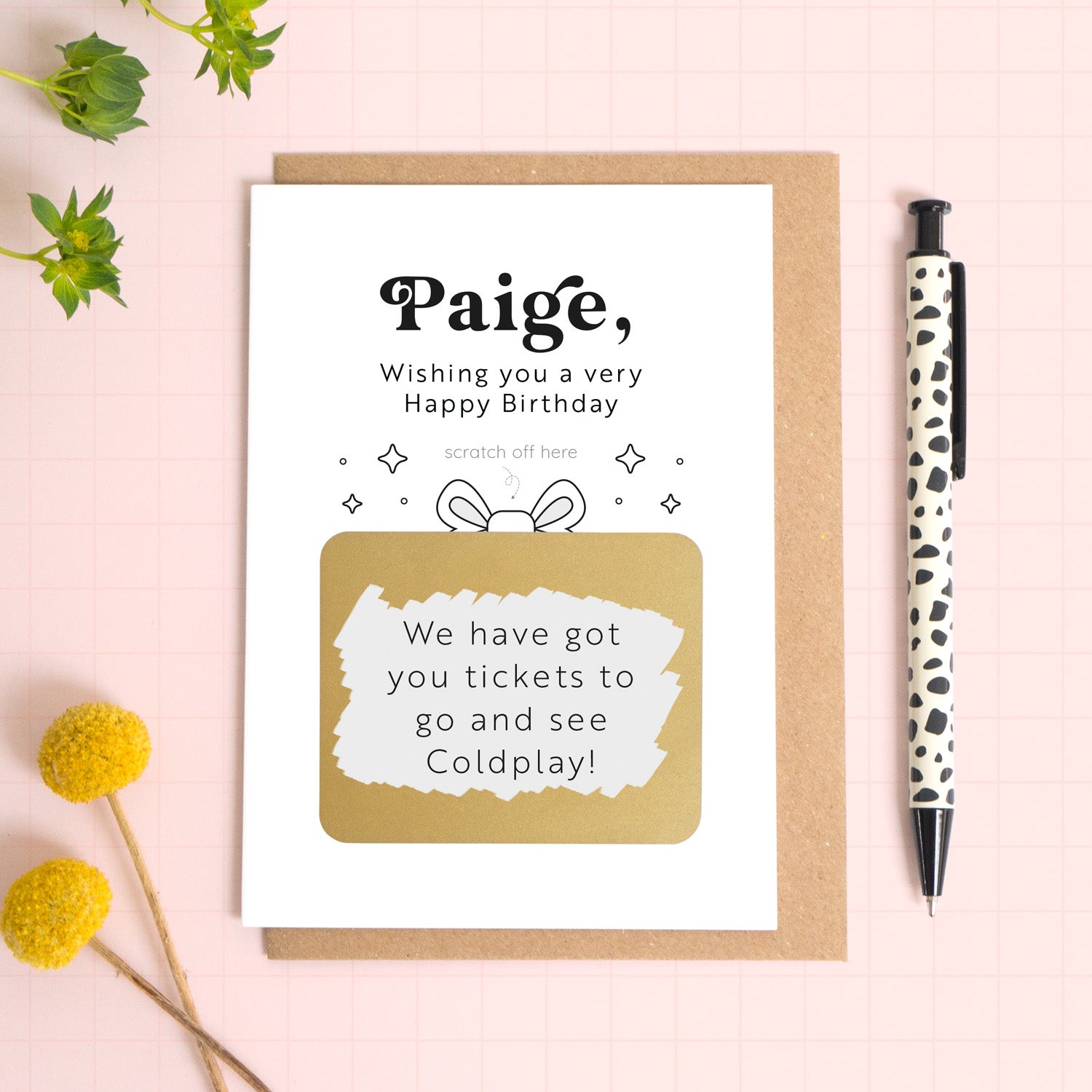 A birthday gift scratch and reveal card where the gold panel has been scratched off to reveal the surprise of gig tickets! The card has been photographed on top of a kraft brown envelope on a pink background. There is foliage and a pen for scale.