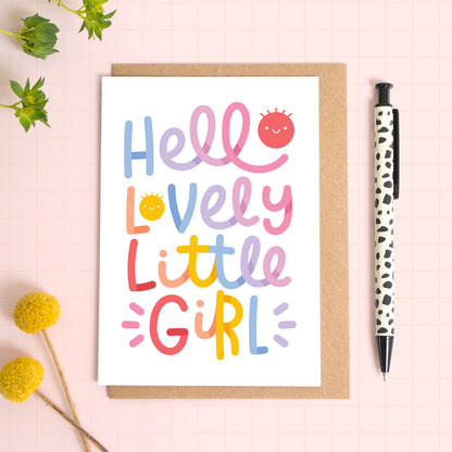 Colourful new baby congratulations greeting card with 'Hello Lovely Little Girl' text on a pink background with a pen and flowers.