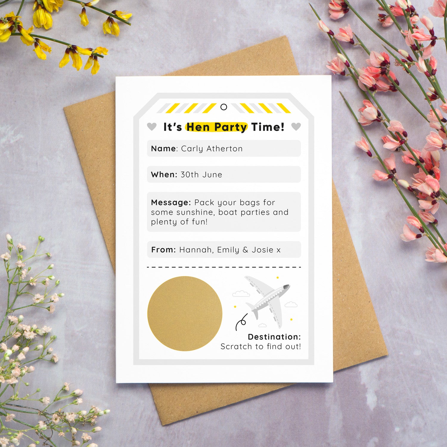 A hen party scratch and reveal card featuring a grey luggage tag and scratch off gold circle that has been photographed on a grey background with yellow, white and pink flowers. The card is lying on a Kraft brown envelope. The scratch off panel has not yet been scratched off to reveal the hen do destination.