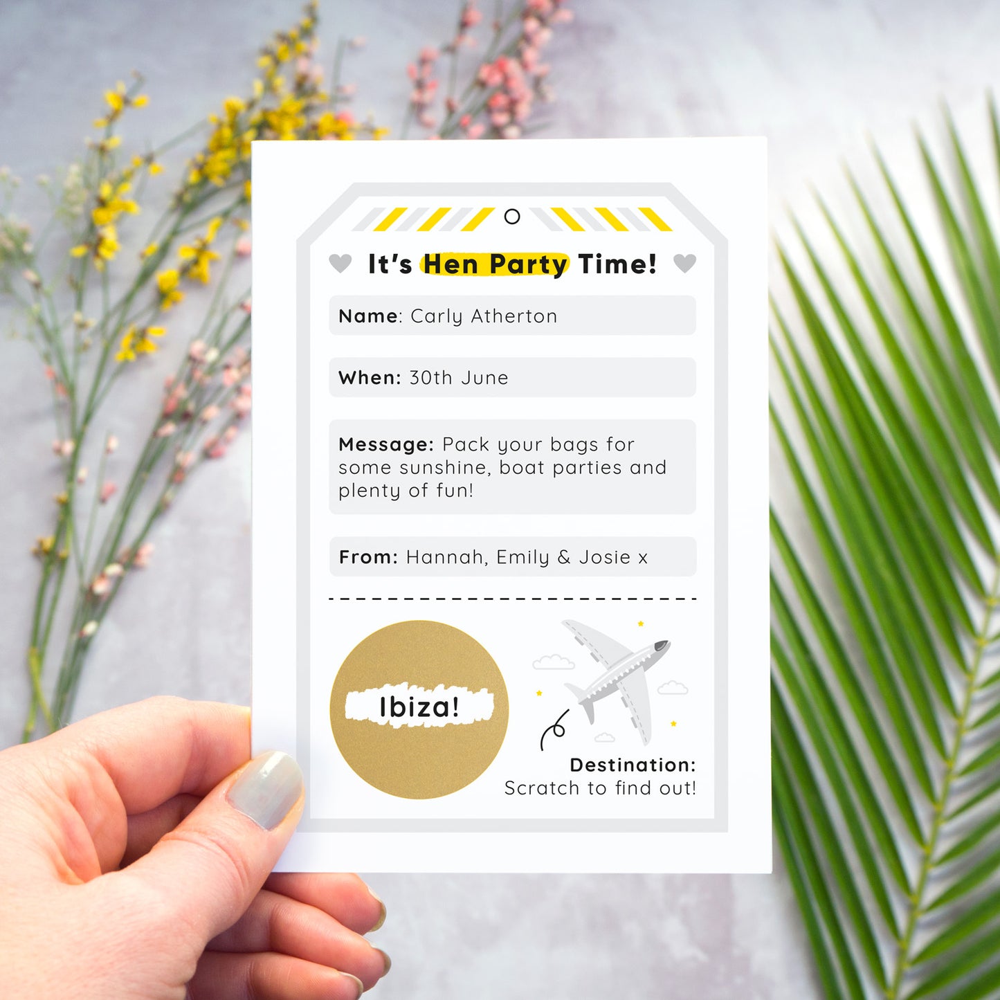 A hen party scratch and reveal card featuring a grey luggage tag and scratch off gold circle that has been photographed being held over a grey background with yellow and pink flowers and green foliage. The scratch panel has been scratched off to reveal the hen do destination.