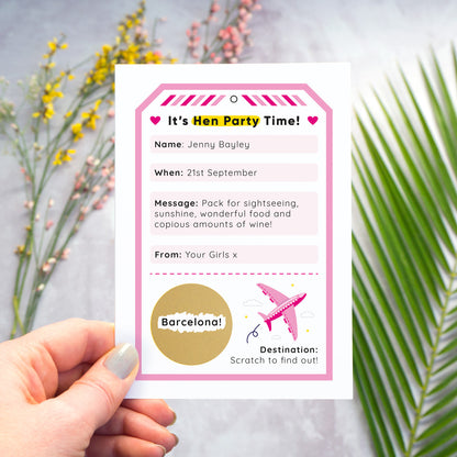 A hen party scratch and reveal card featuring a pink luggage tag and scratch off gold circle that has been photographed being held over a grey background with yellow and pink flowers and green foliage. The scratch panel has been scratched off to reveal the hen do destination.