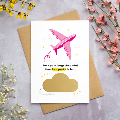 A hen party scratch and reveal card featuring a pink aeroplane and scratch off gold cloud that has been photographed on a grey background with yellow, white and pink flowers. The card is lying on a Kraft brown envelope. The cloud has not yet been scratched off to reveal the hen do destination and date.